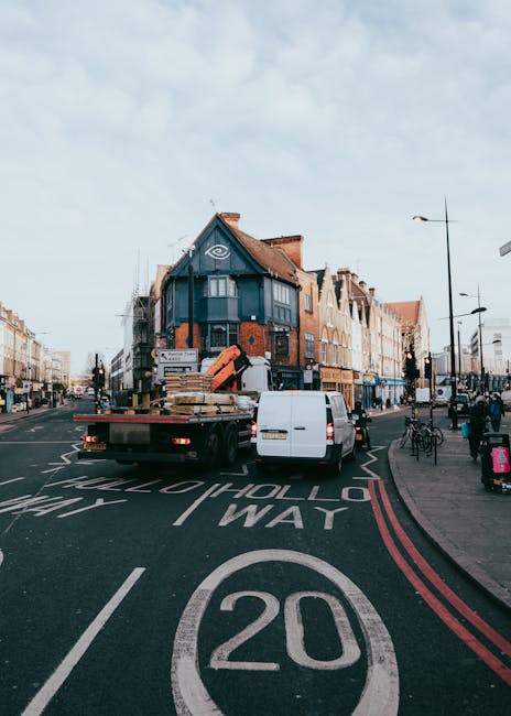 A white flatbed truck parked on a busy urban street during daylight hours, with a large load of wooden pallets and packing materials secured on the truck bed. The vehicle is positioned across the road